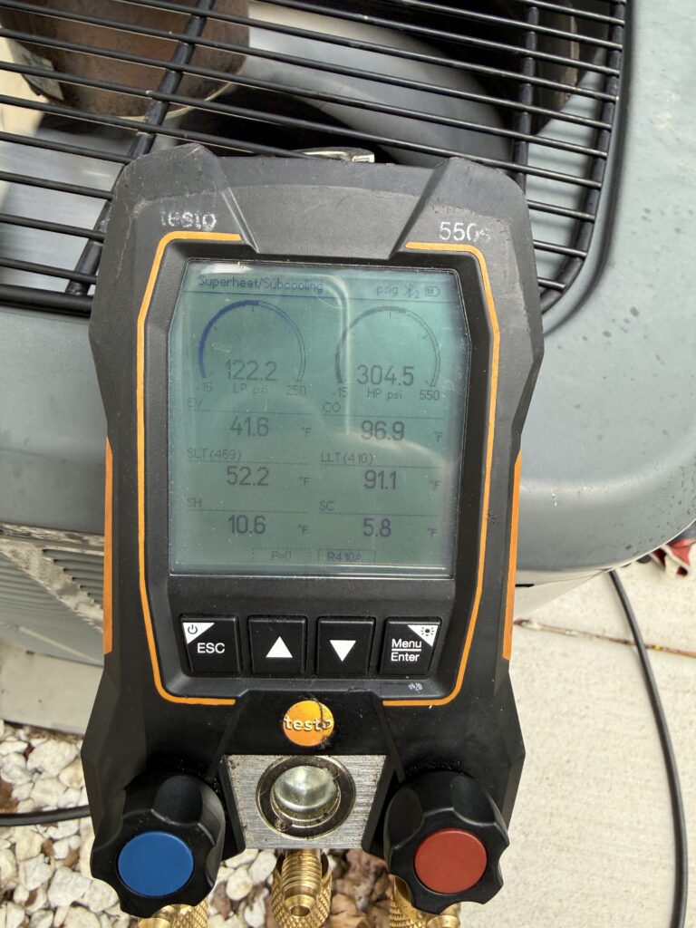 Handheld HVAC diagnostic gauge with blue and red knobs, screen showing pressure readings outdoors.